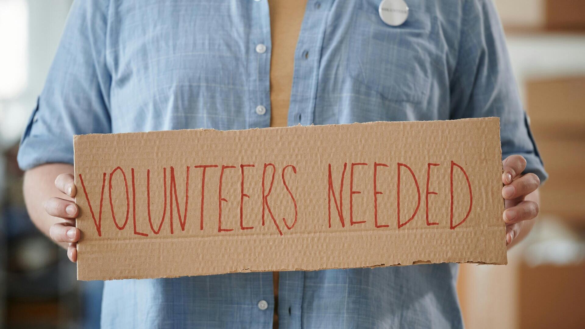 Person holding sign: Volunteers Needed
